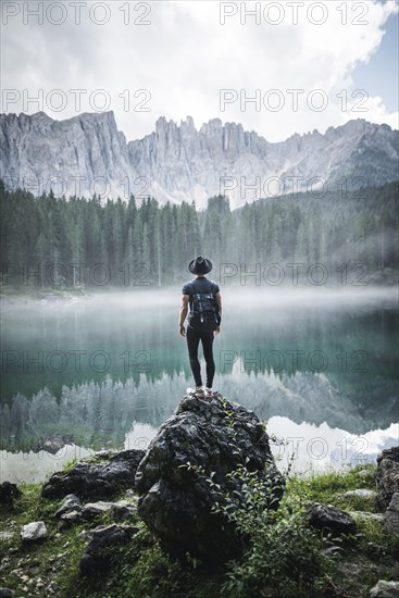 Italy, Carezza, Young man standing on rock at Lago di Carezza in Dolomite Alps at dawn