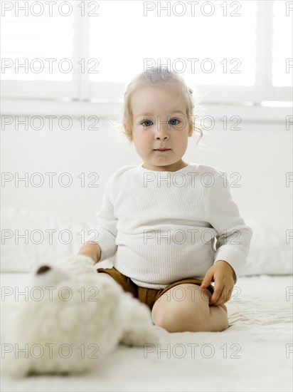 Smiling toddler girl sitting on bed with teddy bear toy