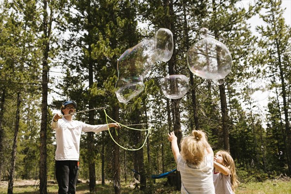 Father making large soap bubbles in forest for two daughters