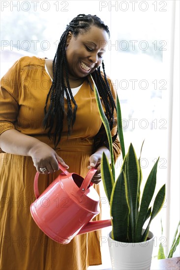 Smiling woman in orange dress watering potted plant at home