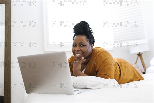 Portrait of smiling woman lying on bed in front of laptop