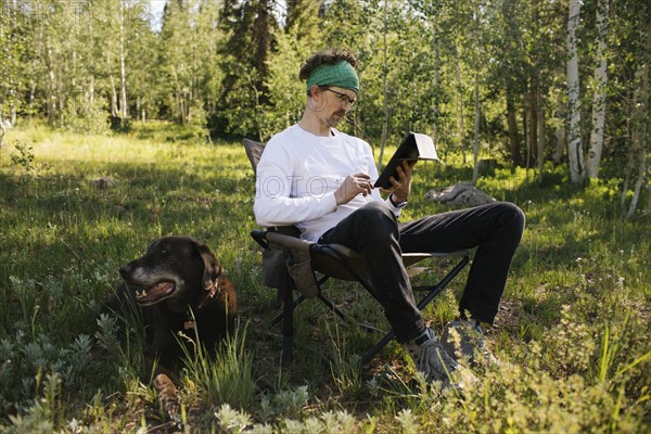 USA, Utah, Uinta National Park, Man with dog sitting in meadow in forest, using tablet