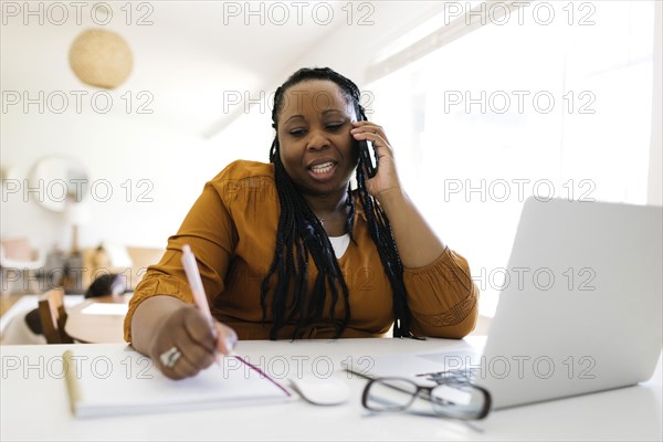 Woman working at desk at home, writing and talking on mobile phone