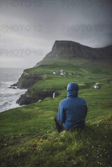 Denmark, Faroe Islands, Gasadalur village, Man sitting and looking at cliffed coast and ocean