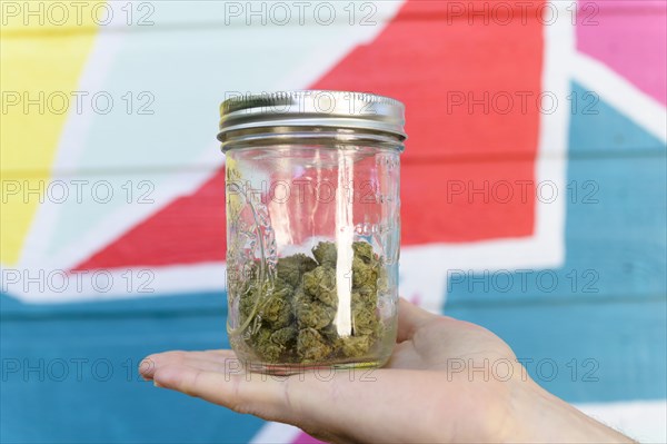 Close-up of mans hand holding jar with cannabis buds in front of colorful mural