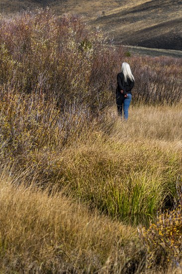 Senior woman with long white hair walking by stream among grass in non urban landscape