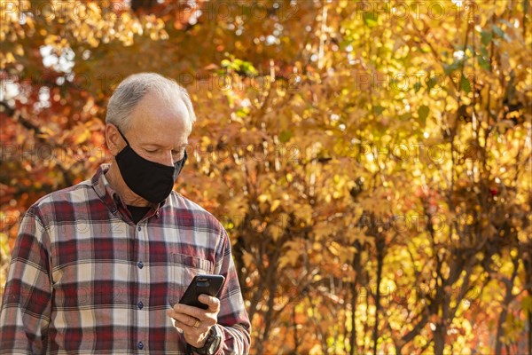 Senior man wearing Covid protective mask using smartphone outdoors in fall