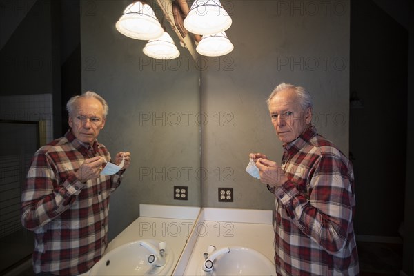 Senior man holding Covid protective mask in front of mirror in bathroom