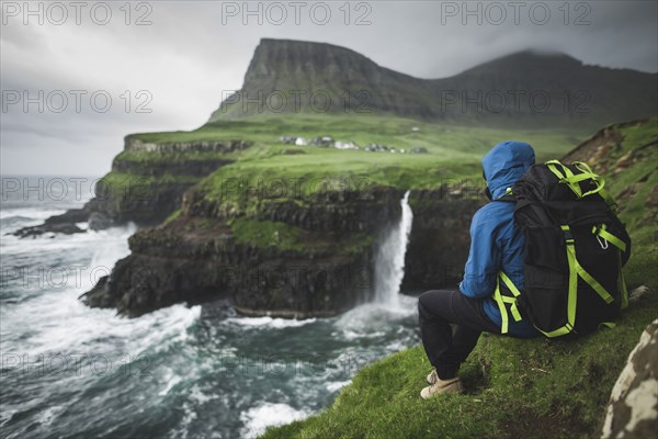 Man with backpack sitting on edge of cliff and looking at Mulafossur Waterfall