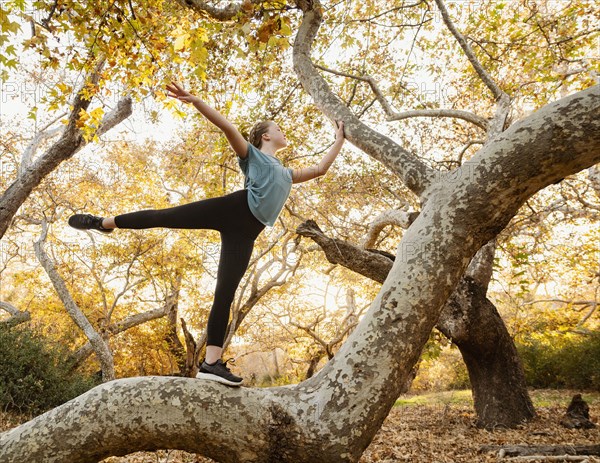 Girl standing on one leg on tree in forest at sunset