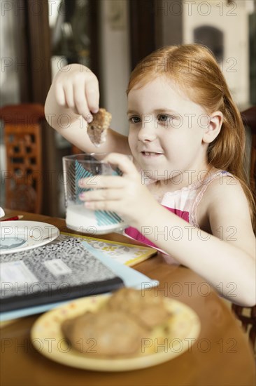 Girl dipping biscuit into glass of milk at dining room table