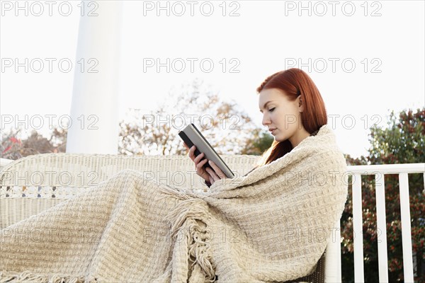 Young woman wrapped in blanket reading digital tablet on porch