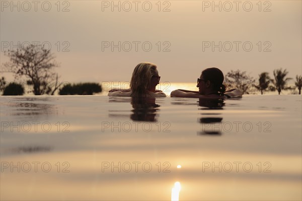 Rear head and shoulder view of two women chatting in rural infinity pool at sunset