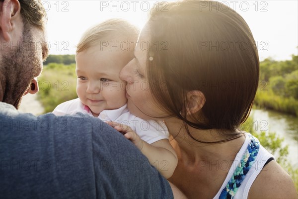 Over shoulder view of mid adult parents kissing baby daughter on riverside