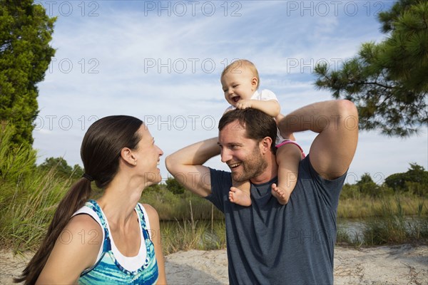 Mid adult parents with baby daughter on mans shoulders by riverside