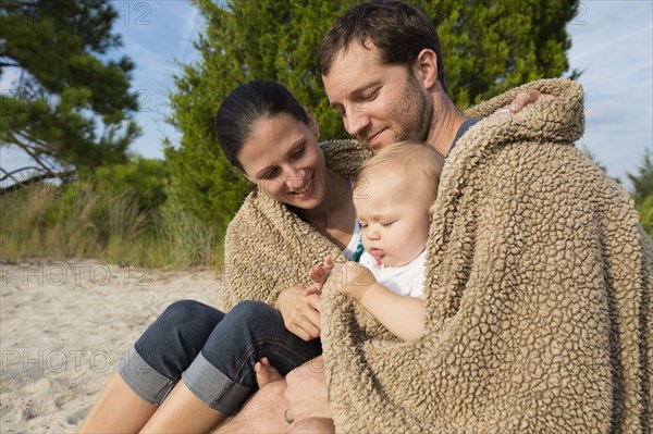 Mid adult parents and baby daughter sitting on sand wrapped in blanket