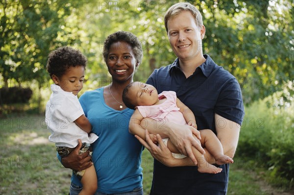 Family outdoors posing for photograph holding preschool boy and baby girl looking at camera smiling