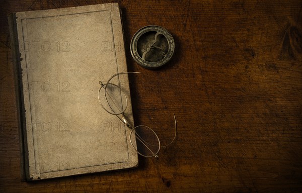 Old book with blank cover resting on old wooden desk top with old eyeglasses and small sundial