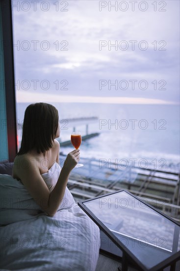 Woman sitting on bed with cocktail in hotel and observing sunrise over sea