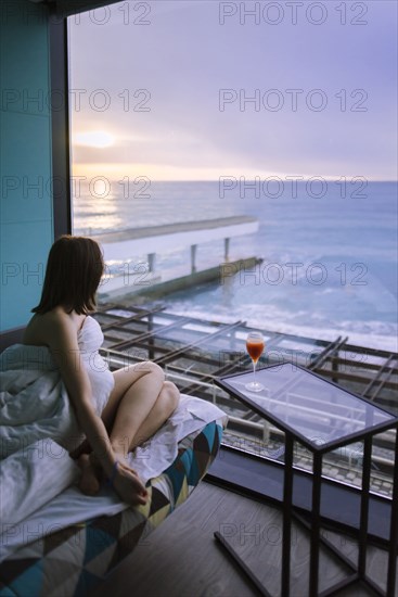 Woman sitting on bed with cocktail in hotel and observing sunrise over sea