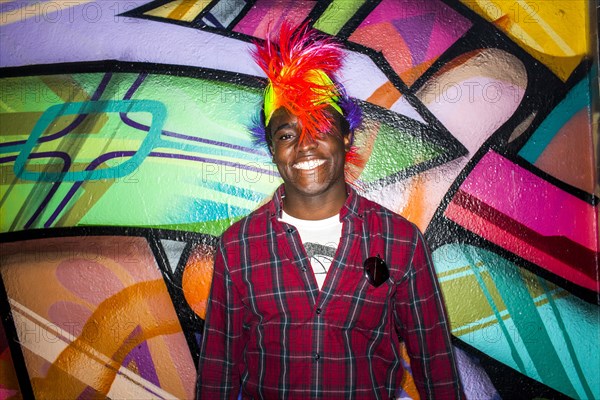 Black man in colorful wig smiling near graffiti wall