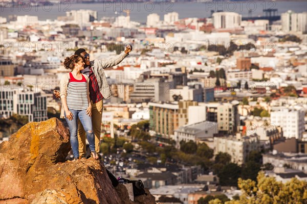 Couple taking cell phone selfie on rock overlooking scenic view of cityscape