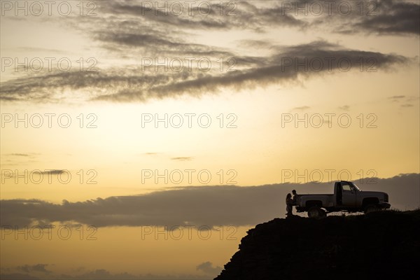Silhouette of people sitting on pickup truck on cliff at sunset