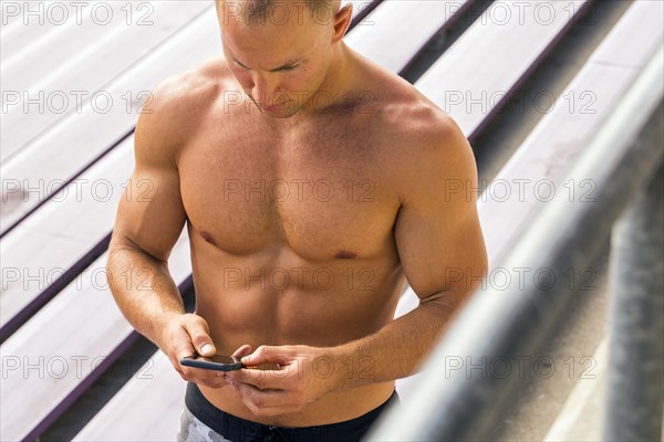 Caucasian man with bare chest standing on bleachers texting on cell phone