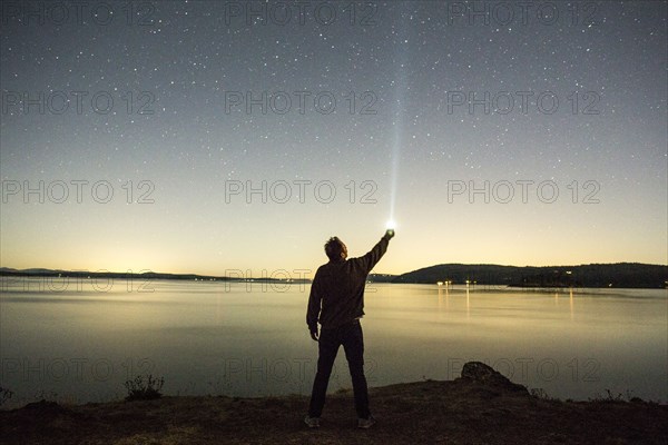 Caucasian man pointing flashlight at night sky near water