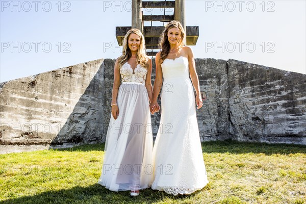 Caucasian brides holding hands in grass near concrete structure