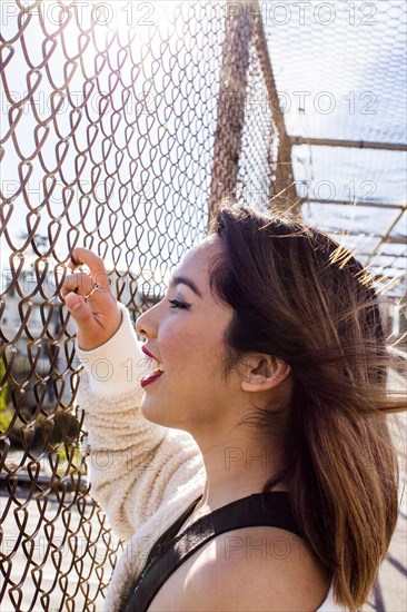 Smiling Asian woman holding chain-link fence on footbridge