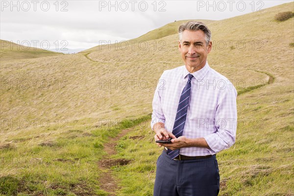 Smiling Caucasian businessman standing in grass holding cell phone