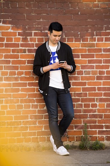 Chinese man leaning on brick wall texting on cell phone