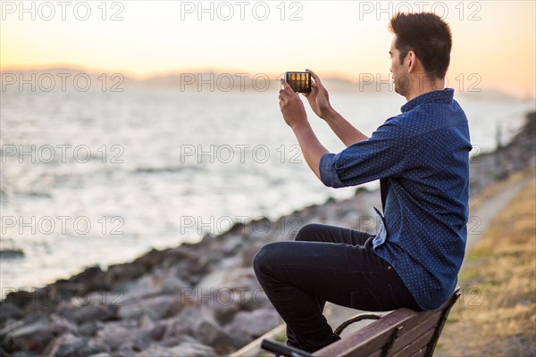 Chinese man sitting on bench photographing ocean with cell phone