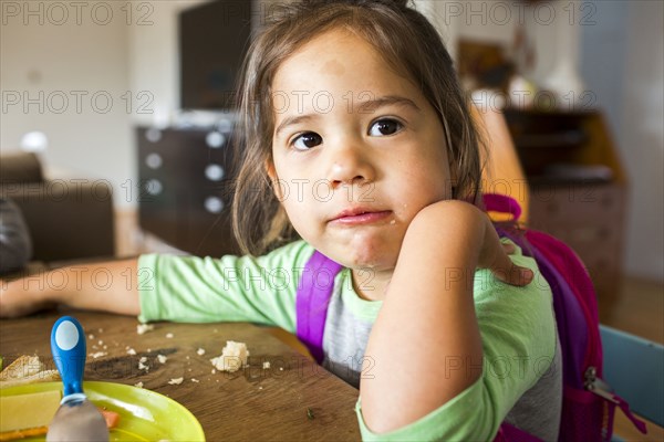 Messy mixed race girl eating food and wearing backpack at table