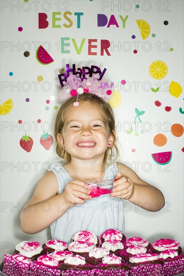 Caucasian girl holding sprinkles over tray of cupcakes for birthday