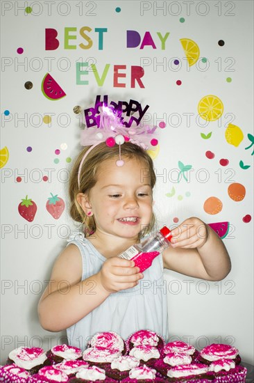 Caucasian girl holding sprinkles over tray of cupcakes for birthday