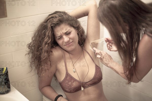Caucasian woman waxing armpit of friend in bathroom