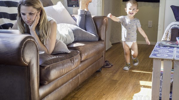 Caucasian baby boy walking past mother on sofa