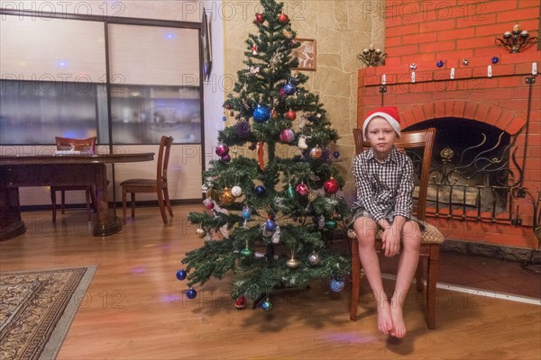 Caucasian boy sitting on chair wearing Santa hat near Christmas tree