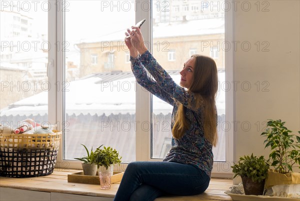 Caucasian woman sitting at window posing for cell phone selfie
