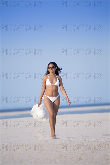 Smiling Mixed Race woman carrying sun hat on beach