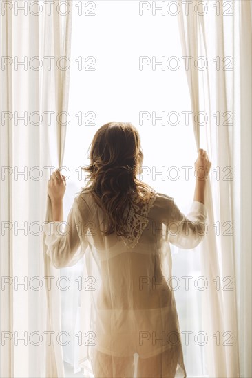 Thai woman wearing sheer clothing opening curtains at window