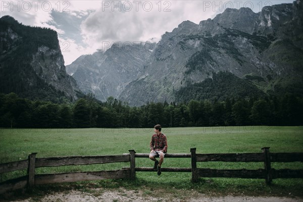 Caucasian man sitting on wooden fence near mountains