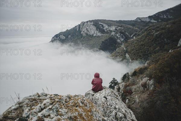 Distant Caucasian woman sitting on rock watching fog on ocean