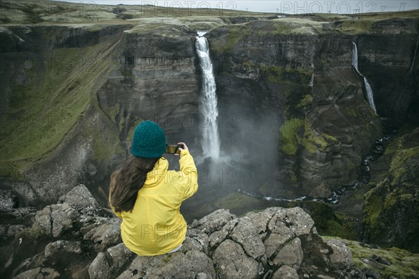 Caucasian woman sitting on cliff photographing waterfall