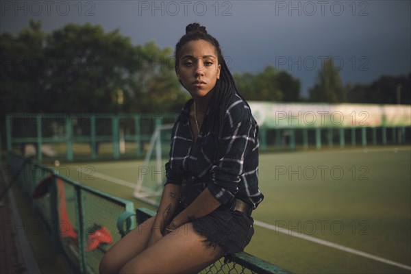 Serious African American woman sitting on fence at sports field