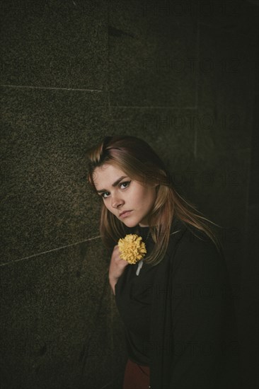Serious Caucasian woman leaning on wall holding flower