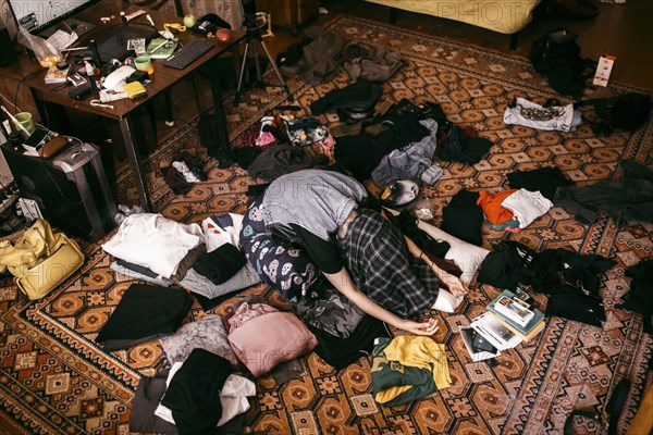 Caucasian man sitting on messy floor with shirt covering head