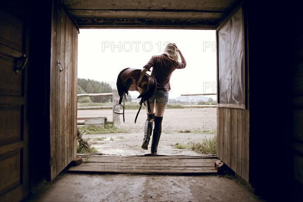 Caucasian woman carrying horse saddle in barn doorway